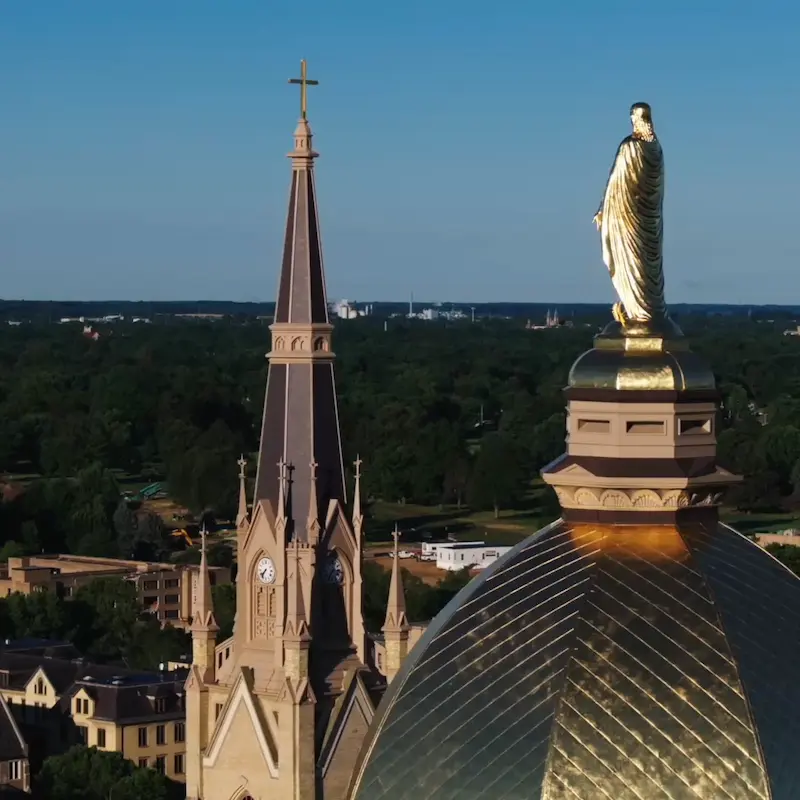 An aerial view of the Golden Dome on the Main Building at the University of Notre Dame, with the Sacred Heart Basilica spire beside it, against a blue sky.